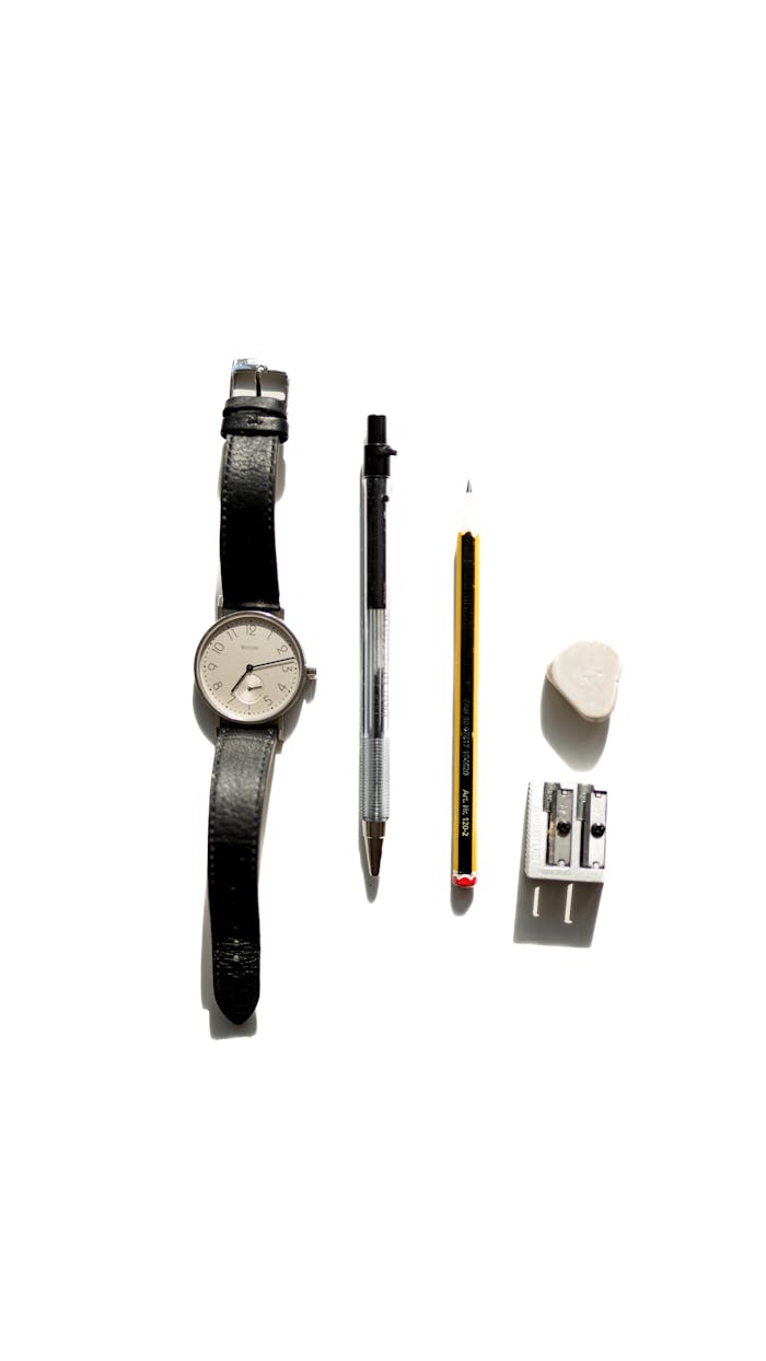 A minimalist still life arrangement of a watch, pen, pencil, eraser, and sharpener on a white surface.